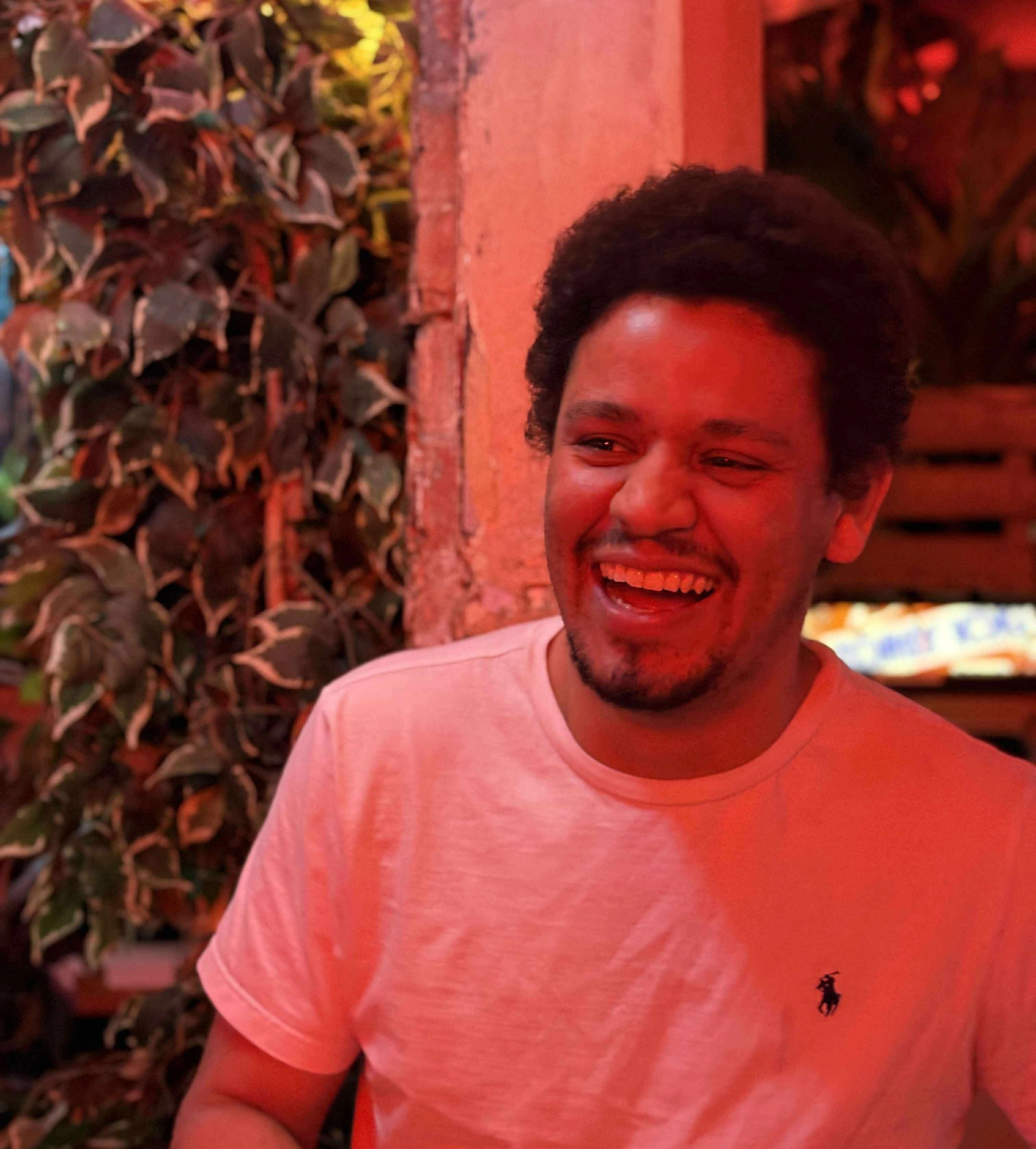 A portrait of a laughing man with short curly dark hair and a goatee, wearing a light T-shirt, in front of leafy plants under warm red light.