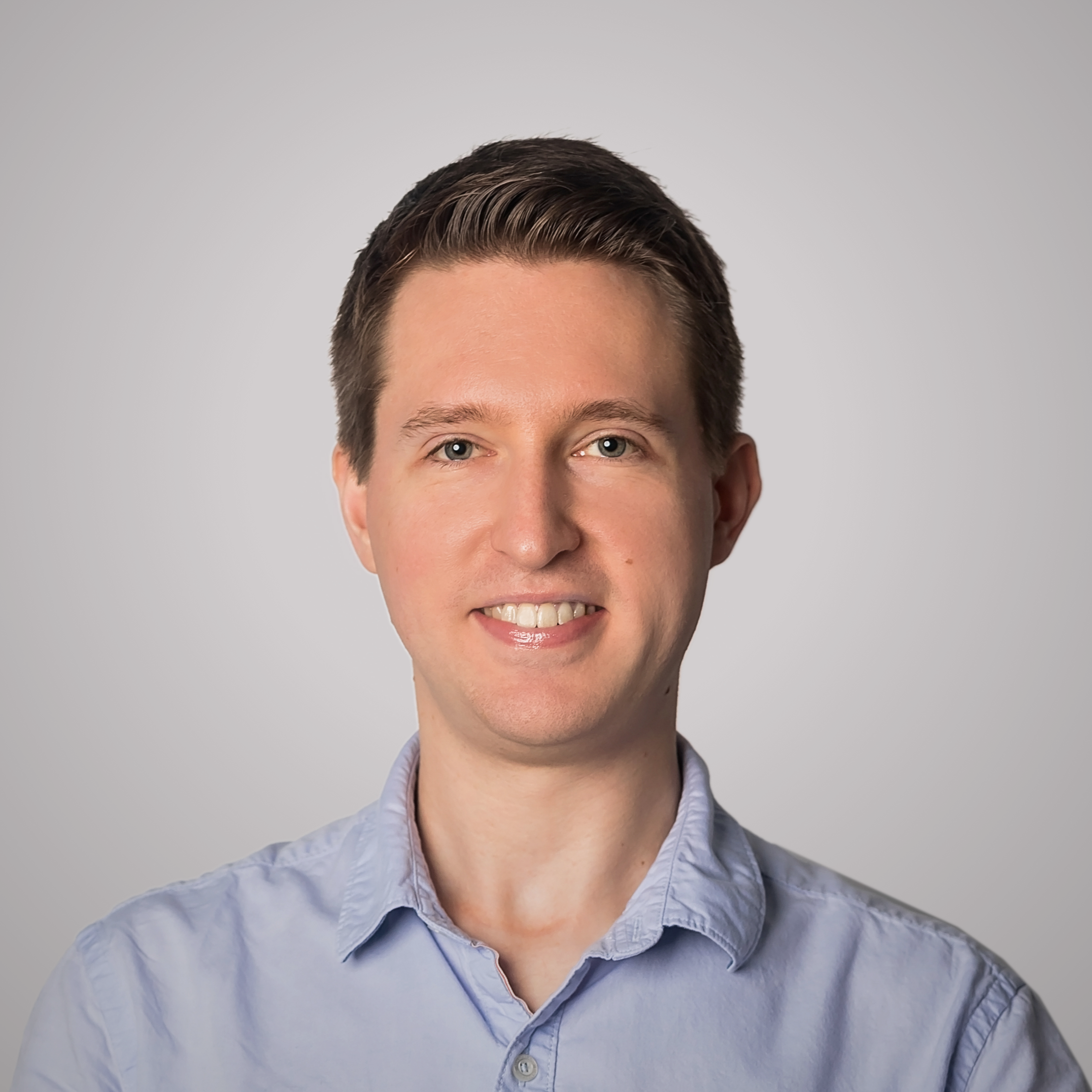 A portrait of a smiling man with short brown hair, wearing a light-blue button-up shirt, against a neutral grey background.